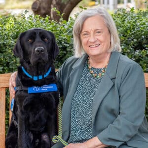 a smiling woman on a bench sitting next to a yellow lab service dog