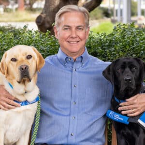 a smiling man sits next to a black lab service dog on a bench