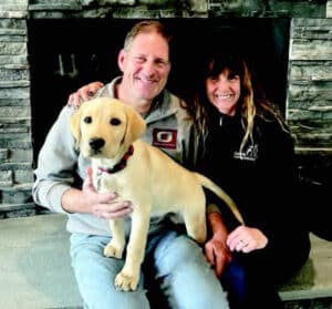 A man and a woman sitting on a stone hearth with a tan Labrador puppy in the man's lap; the background features a stone fireplace.