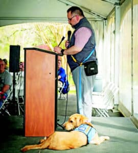 A man stands at a podium with a microphone, wearing a vest and plaid shirt, while a golden retriever service dog lies calmly on the floor beside him in a tented environment with seated audience members in the background.