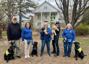 A group of four adults standing outdoors with three black Labrador Retrievers, all wearing yellow training vests, and two puppies in their arms. The adults are dressed in blue shirts and are positioned in front of a house surrounded by bare trees and grass.
