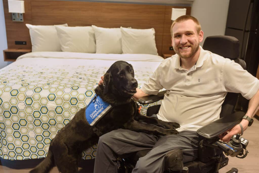 A man in a wheelchair smiles while sitting on a bed, with an assistive dog resting its front paws on his lap. The dog is wearing a blue vest that says 'Canine Companions'. The background shows a neatly made bed with a geometric-patterned duvet and pillows.