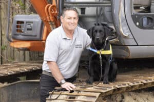 A man kneels beside a black Labrador retriever on the tracks of a construction vehicle, with the dog wearing a yellow vest labeled "Canine Companions." The man is smiling and wearing a grey polo shirt.