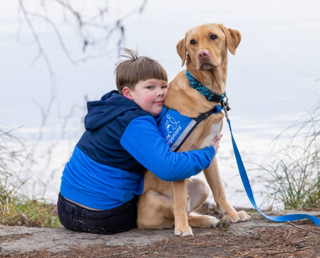 A child wearing a blue jacket sits on the ground, hugging a yellow Labrador retriever, which is wearing a harness. The background features a calm body of water and some grass, creating a serene outdoor setting.