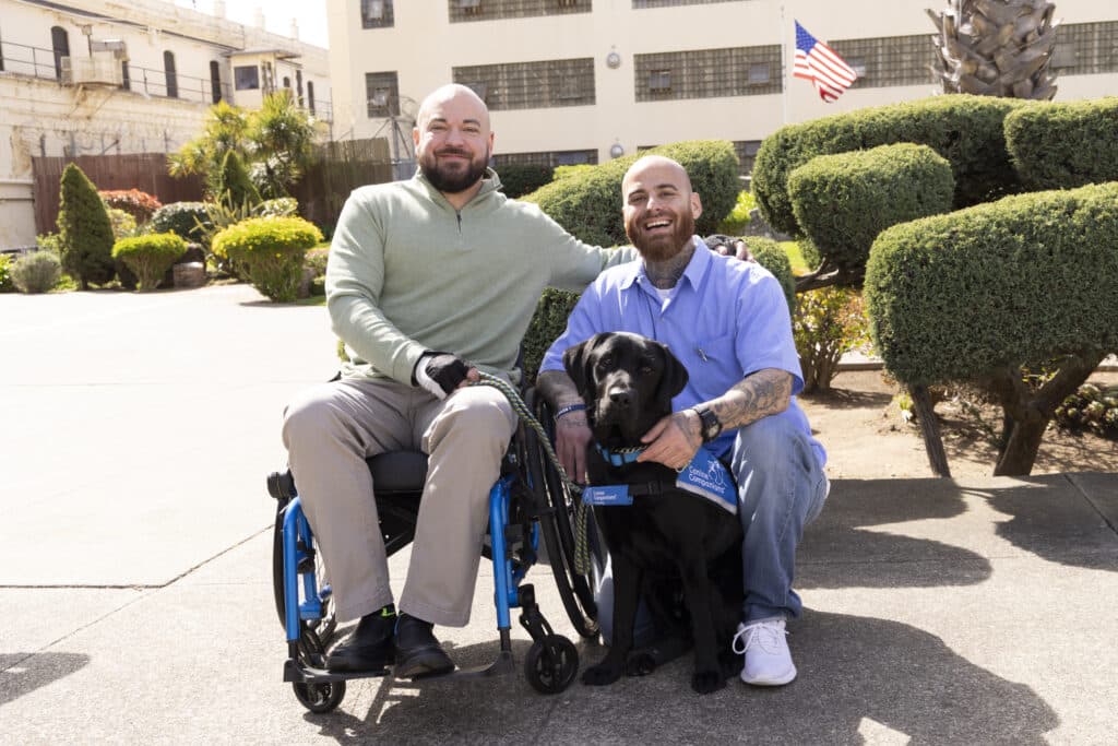 Two men smile while sitting outside; a black service dog sits between them. The background features landscaped bushes and a building with barred windows.