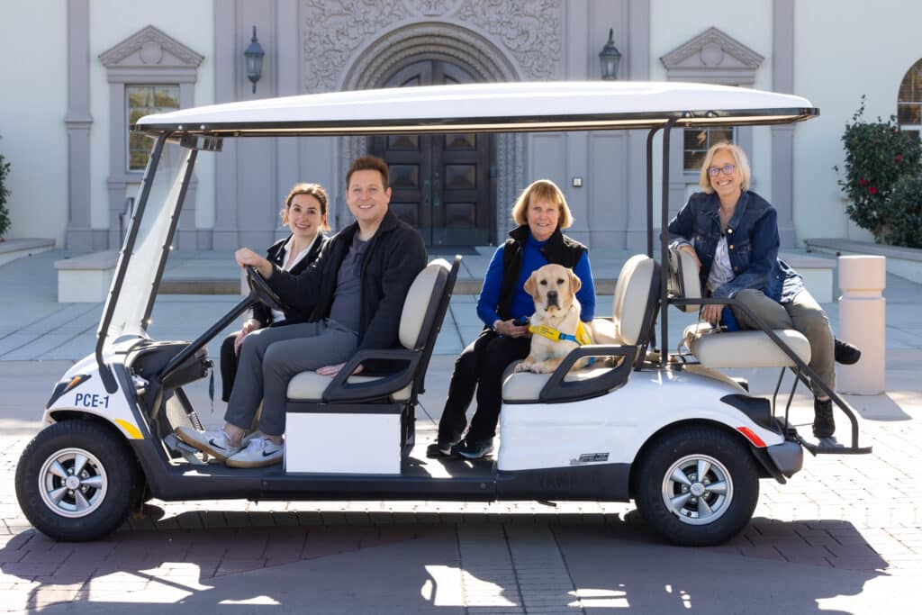 A group of people ride on a golf cart with a puppy in a yellow vest.