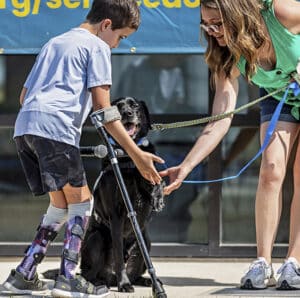 A young boy in leg braces and using crutches shakes hands with a black service dog while a woman holds the dog's leash.