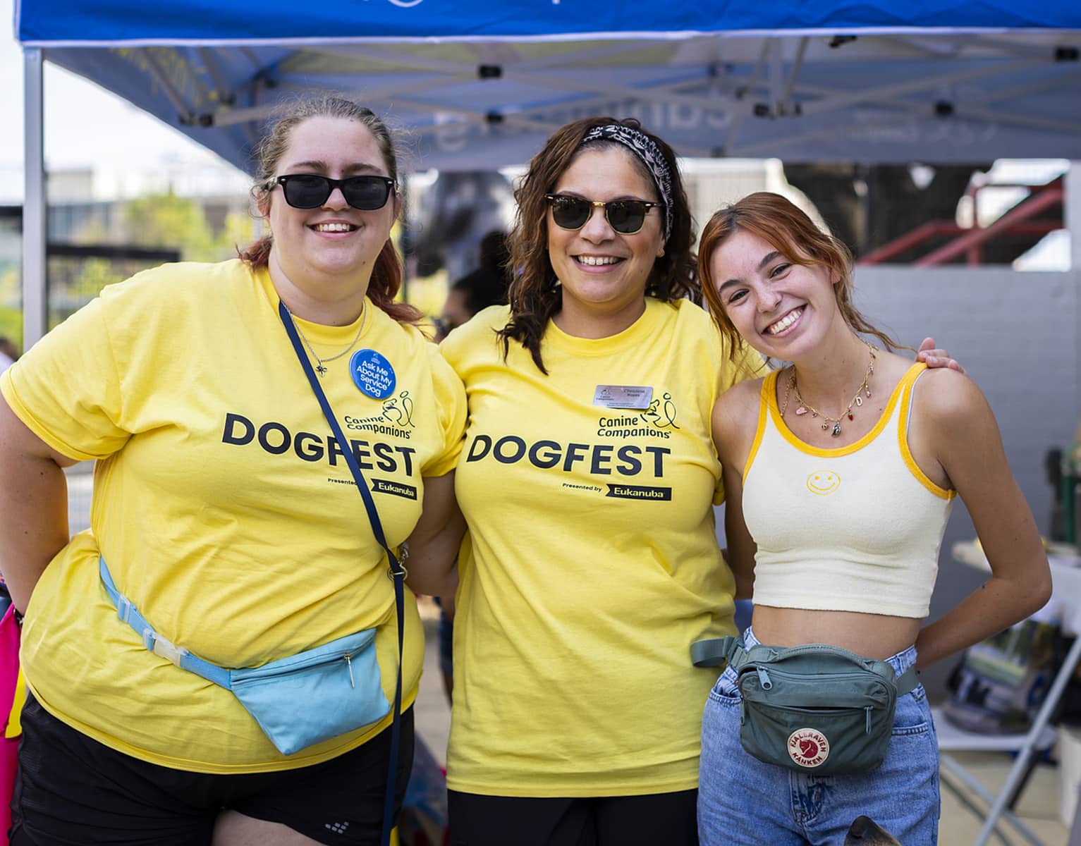 Three people pose together at an outdoor event, all wearing yellow shirts featuring 'DogFest' and 'Canine Companions.' One person wears sunglasses and has a blue fanny pack, while another has a headband and a name tag. The third person wears a light-colored tank top and has a green fanny pack, appearing cheerful in a festive setting.