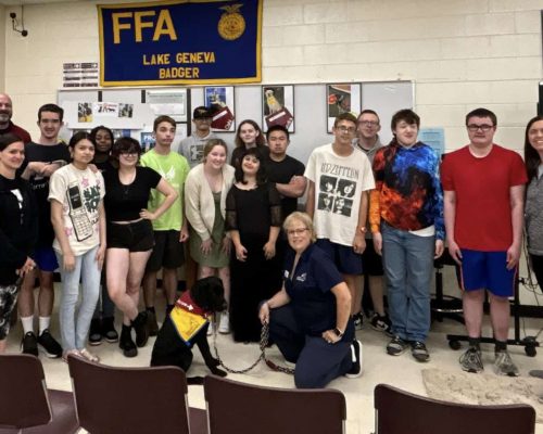 FFA_Minicrop2 Group of people posing in a room with an FFA Lake Geneva Badger banner, featuring a seated black dog wearing a yellow and red vest.