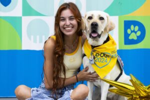 A young woman with long, wavy hair smiles while sitting next to her Labrador retriever service dog wearing a yellow bandana that reads 'DOG FEST 2024'. They are posing in front of a colorful background featuring geometric shapes.