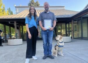 Two people stand outside a building, with the man on the right holding a framed document and a Labrador service dog sitting by his side.