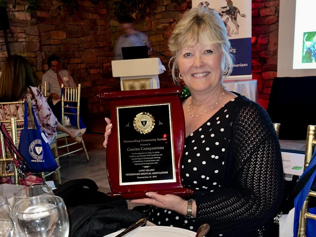 A woman in a black and white polka dot dress sits at a banquet table, holding a large plaque and smiling at the camera.