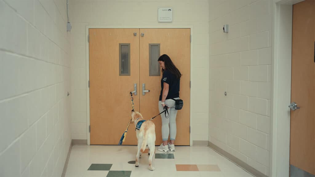 A woman holding the leash of a service dog stands in front of double wooden doors in a hallway.