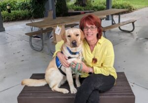 A woman with short, red hair and glasses is sitting on a wooden bench, smiling as she poses with a yellow Labrador retriever wearing a blue vest. The background features a park setting with a picnic table and greenery.