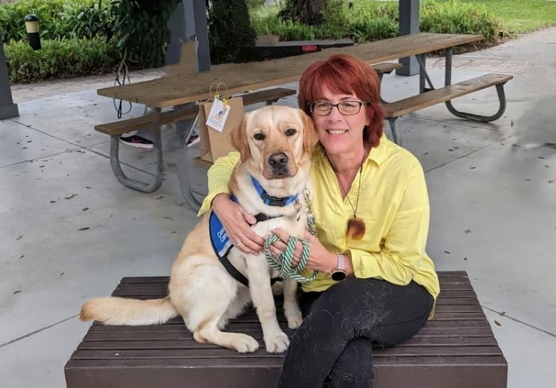 A woman with short, red hair and glasses is sitting on a wooden bench, smiling as she poses with a yellow Labrador retriever wearing a blue vest. The background features a park setting with a picnic table and greenery.