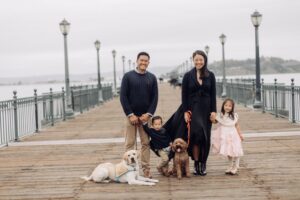 A family of four standing on a wooden pier, with a yellow Labrador Retriever and a brown dog. The father is in a navy sweater, the mother is wearing a black dress, and two children are present: a boy in a navy sweater and a girl in a pink dress. The background shows lanterns and a view of water.