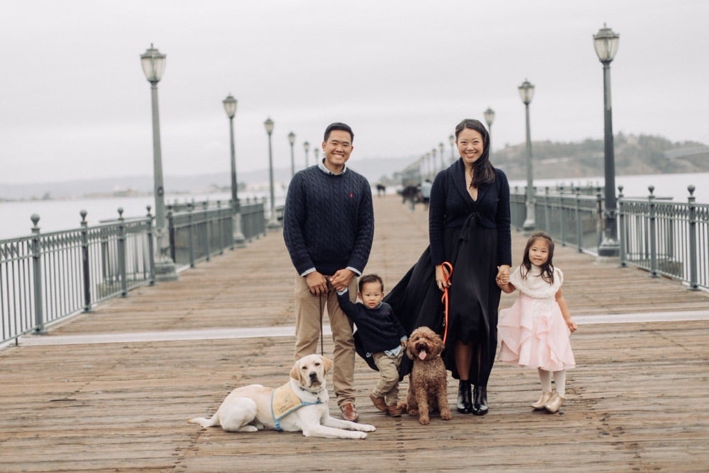 A family of four standing on a wooden pier, with a yellow Labrador Retriever and a brown dog. The father is in a navy sweater, the mother is wearing a black dress, and two children are present: a boy in a navy sweater and a girl in a pink dress. The background shows lanterns and a view of water.