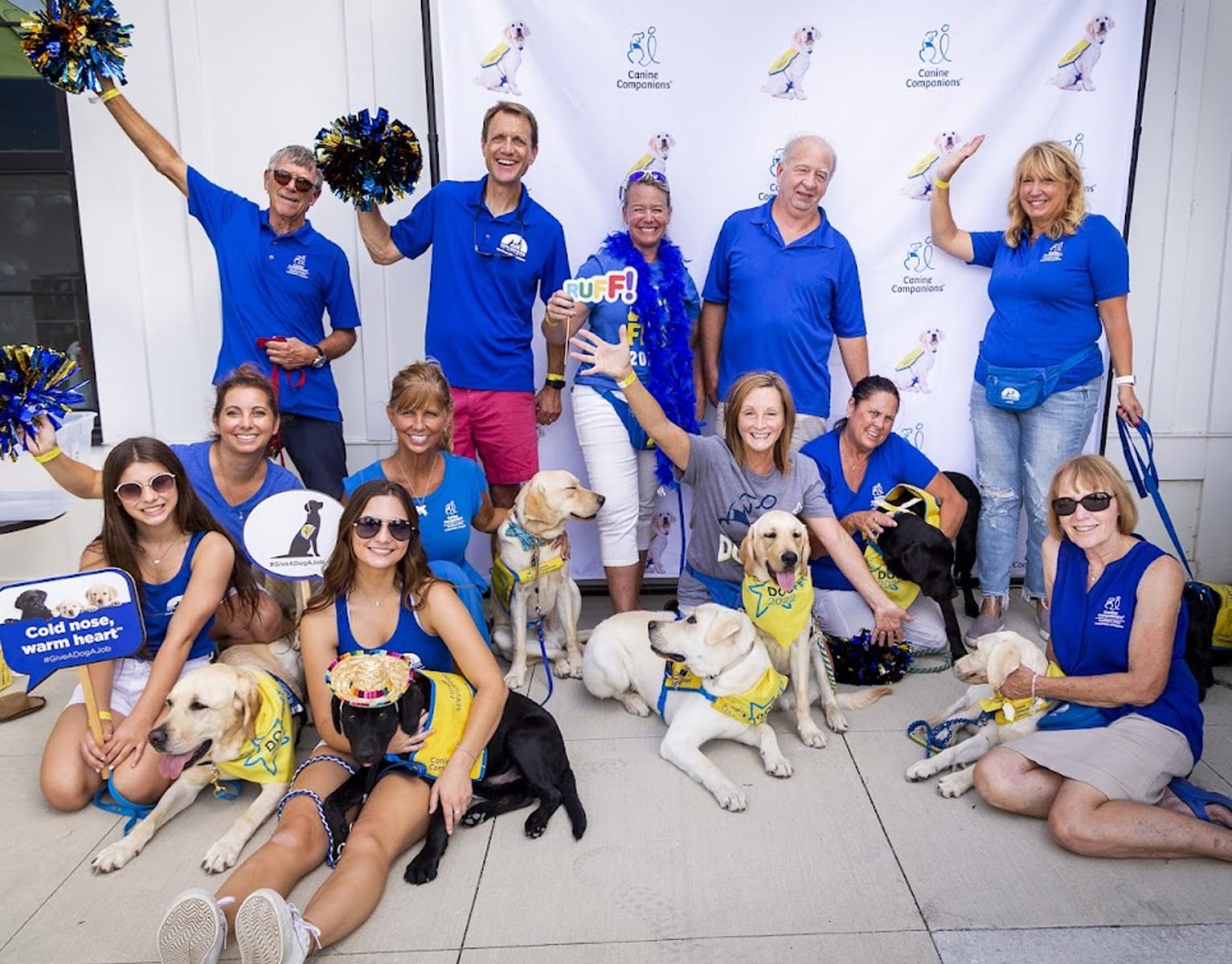 A group of people wearing blue shirts, some holding pom-poms, pose cheerfully with several dogs in yellow bandanas. They celebrate in front of a backdrop with logos related to a dog charity event. Signs with messages about dog adoption are visible.