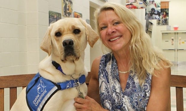Woman sitting next to a yellow Labrador retriever wearing a blue "Canine Companions" vest.