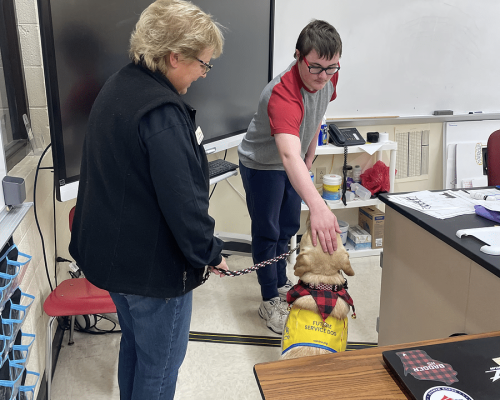 ffa_minicrop4 A person pets a future service dog that is wearing a yellow vest, while another person holds the dog's leash in a classroom.