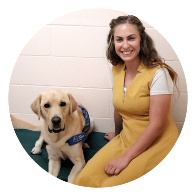 A woman smiling and sitting next to a Labrador wearing a blue service dog vest.