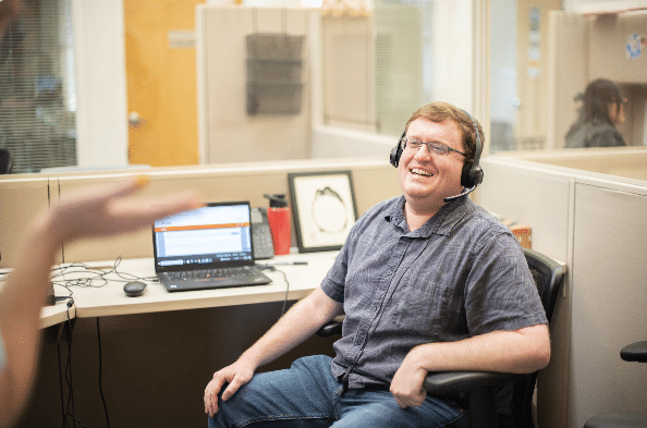 Man sitting at a desk in an office, wearing a headset, and smiling.