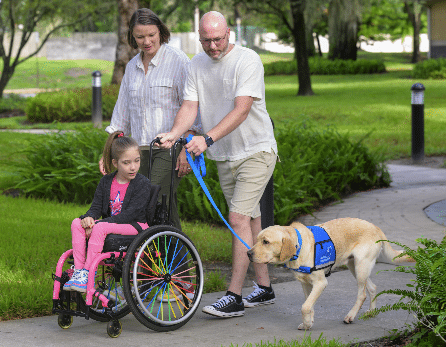A young girl in a wheelchair being pushed by a man, accompanied by a woman and a service dog on a leash, walking on a path in a park.